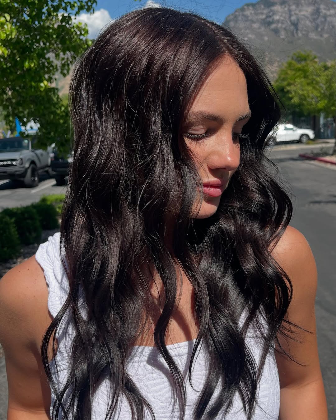 Close-up of a woman with long, shiny dark brown hair styled in soft waves, showcasing a beautiful dark summer hair idea. Her hair reflects sunlight, highlighting subtle natural undertones. Perfect for elegant summer styling.