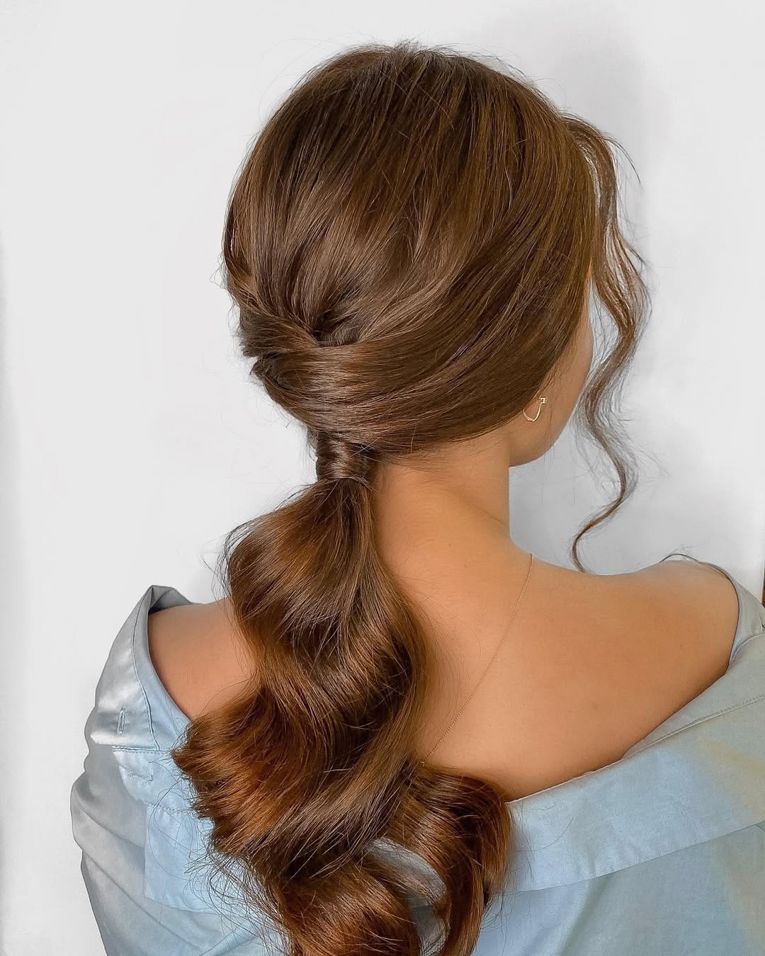 Close-up of a woman with a sleek, high ponytail, styled with two face-framing tendrils, perfect for easy summer hairstyles 2026. Her hair is black and glossy against a softly blurred outdoor background.