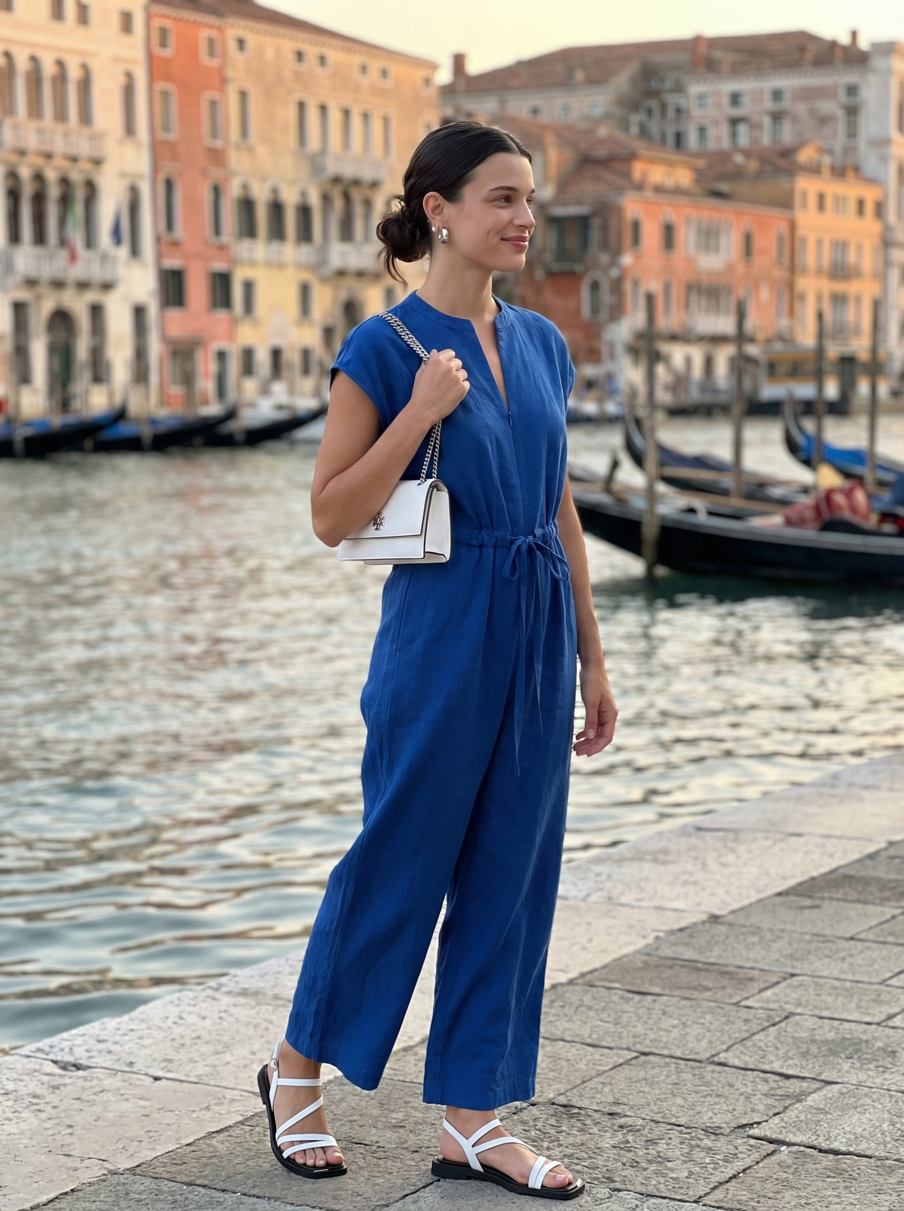 Chic Europe summer outfits for a Venetian canal cruise, featuring an ocean blue wide-leg jumpsuit, white leather flat sandals, a white structured shoulder bag, and delicate silver hoop earrings.