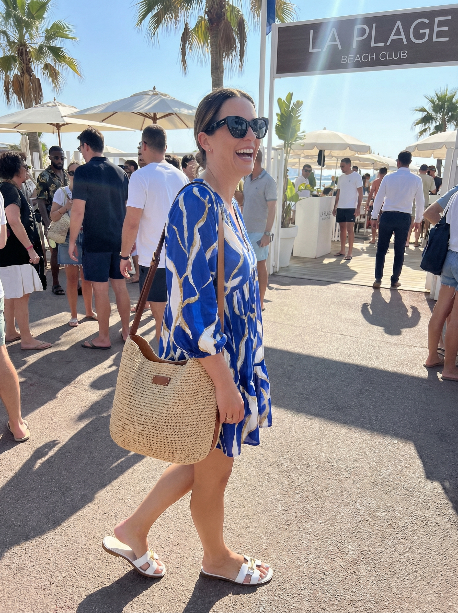 A playful European summer outfit for 2026 with a cobalt blue abstract print mini shirt dress, gold metallic slide sandals, a woven raffia crossbody bag, and oversized cat-eye sunglasses, perfect for a beach club.