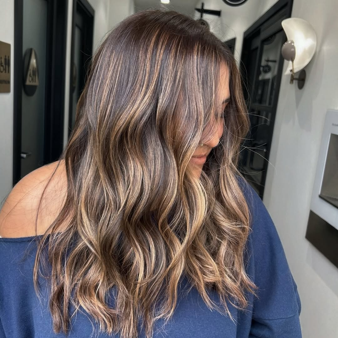 Woman with long, wavy blonde hair with subtle caramel highlights, showcasing a perfect soft summer hair color 2026. She's wearing a delicate white lace top, styled casually.