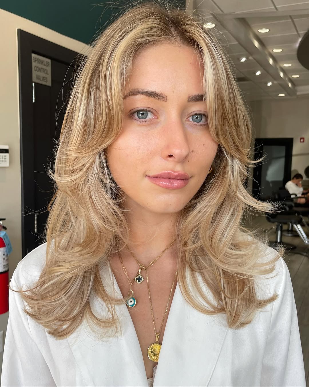 A woman with a chic, sun-kissed soft summer hair color 2026, posing in a stylish fall outfit. She's wearing a cream knitted sweater, dark wash jeans, and ankle boots, perfect for a casual autumn day.