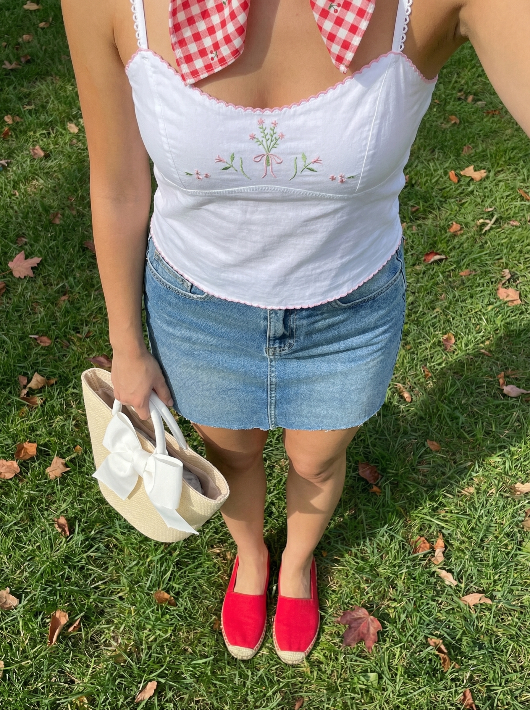 Playful park picnic spring summer outfits with a light wash denim mini skirt, white embroidered crop top, red espadrille flats, a white straw basket bag, and a gingham hair scarf.