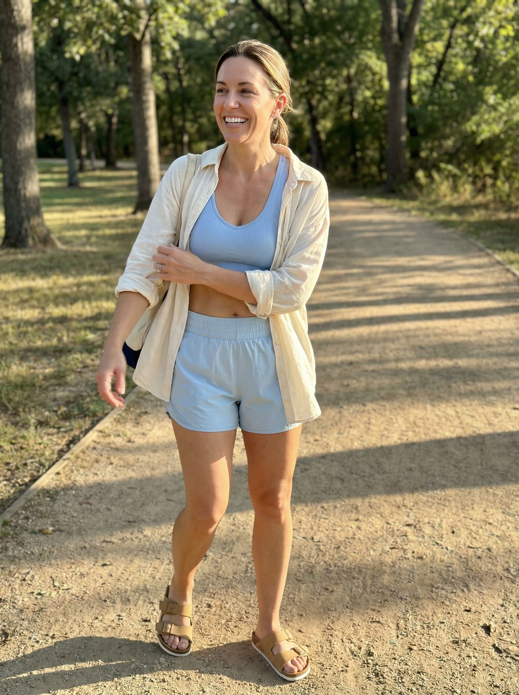 Post-workout summer athleisure outfit with a sky blue sports bra, white athletic shorts, an oversized linen shirt, and tan slide sandals, perfect for casual days.