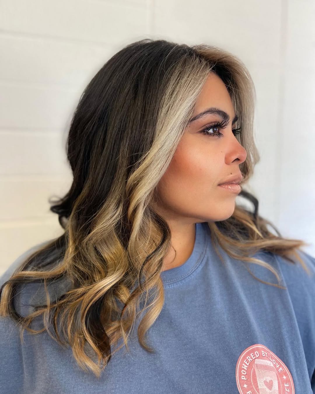 Close-up of a woman with long, wavy blonde hair styled in a half-updo with loose face-framing tendrils, perfect for summer hair ideas 2026. She wears a casual white top, highlighting the effortless elegance of the hairstyle.