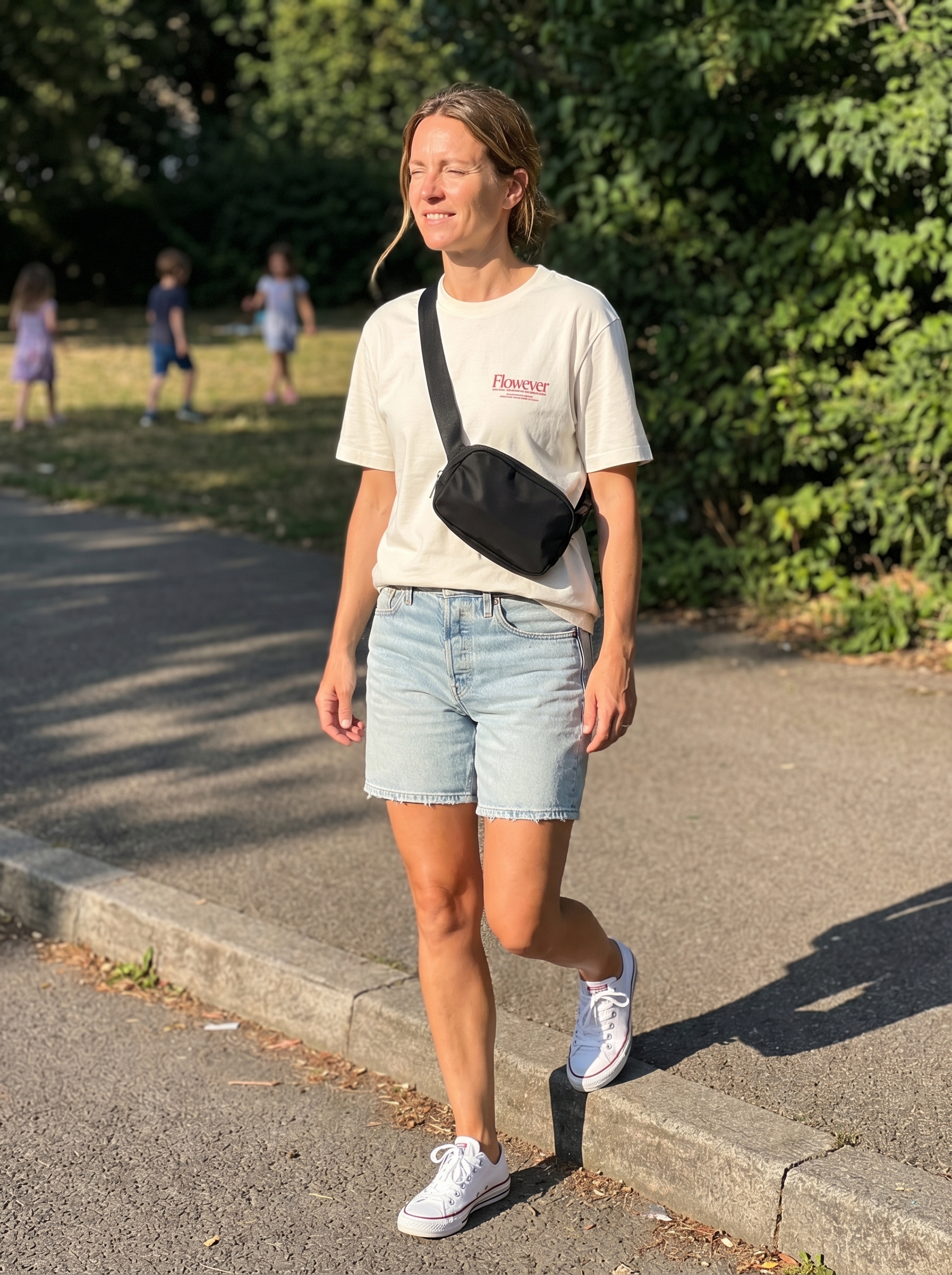 Casual park playdate summer mom outfit with light wash denim shorts, a graphic tee, white sneakers, and a black belt bag. Comfortable and youthful style for 2026.