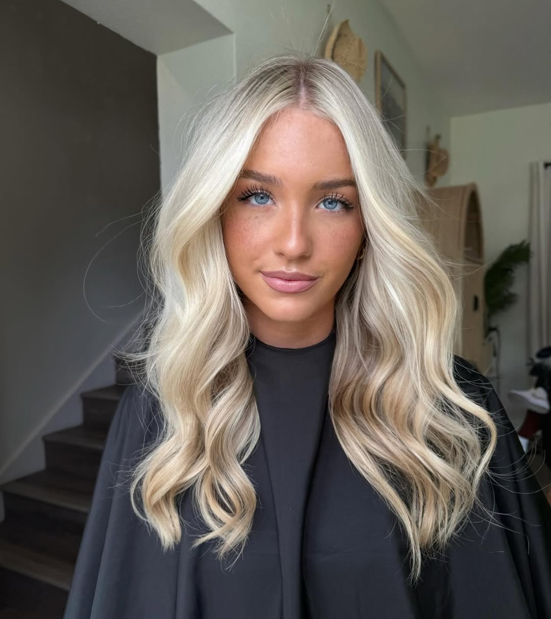 A woman with radiant blonde hair inspo summer 2026 smiles, wearing a white off-the-shoulder top and delicate gold jewelry, perfect for a sunny day.