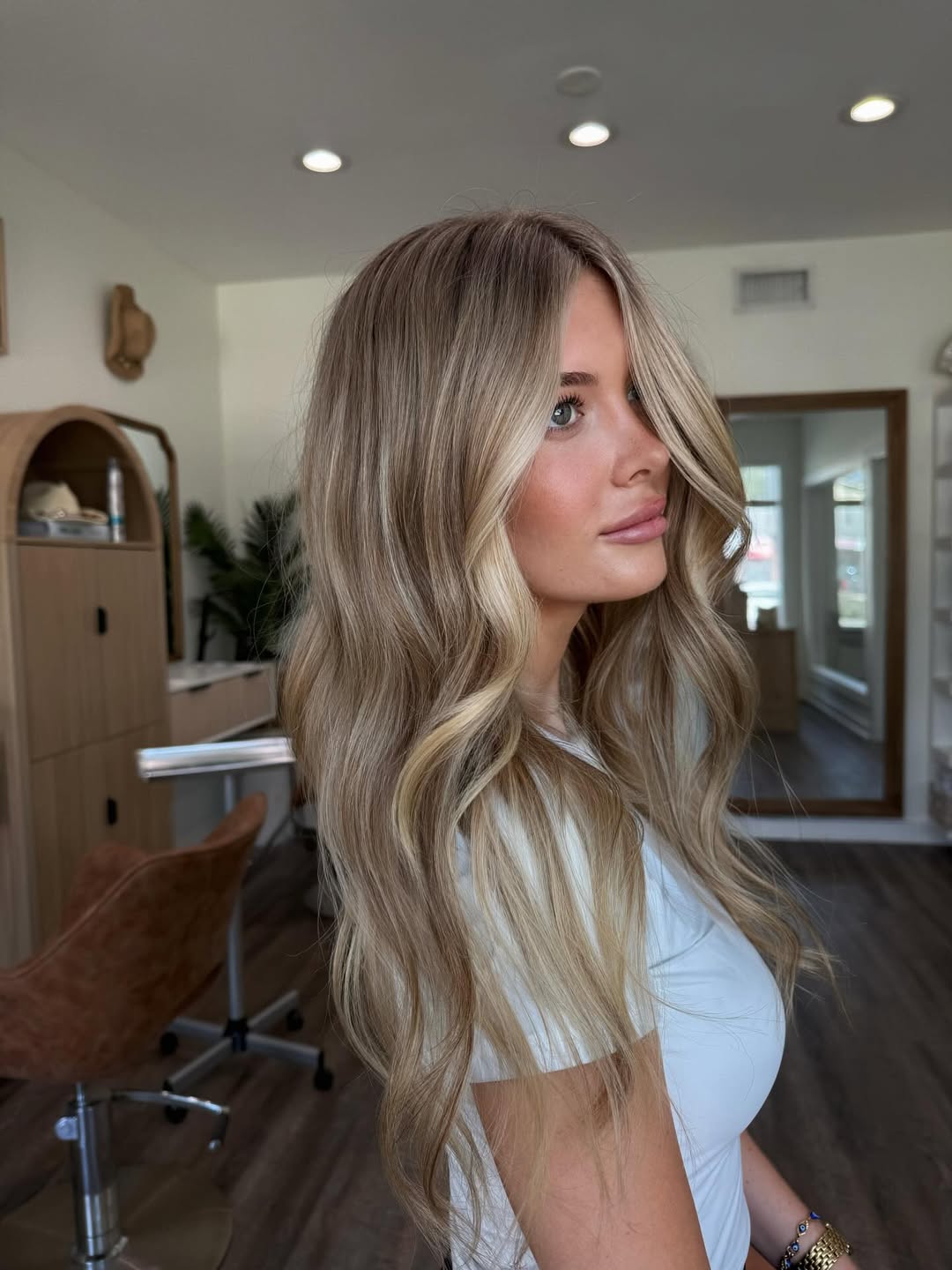 A woman with radiant blonde hair, styled in loose waves, enjoys a sunny day. She wears a flowy white sundress, perfect for blonde hair inspo summer 2026. Her hair catches the light beautifully, highlighting her relaxed and chic aesthetic for warm weather.