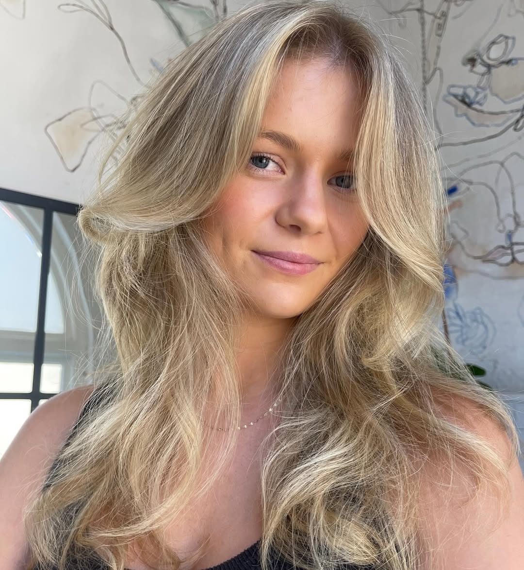 Beautiful woman with long, shiny brown hair styled in soft, natural waves, perfect for cute summer hair ideas 2026. She wears a simple white top, showcasing a fresh, effortless look.