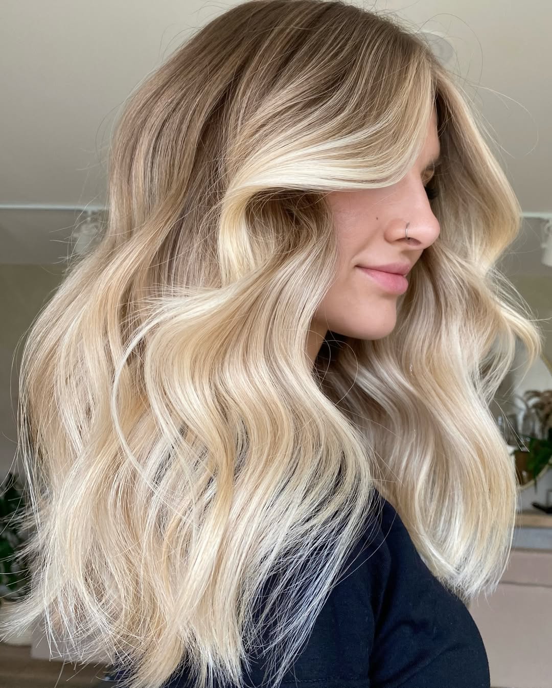 Woman with long, wavy soft summer blonde hair 2026, styled casually, wearing a white off-the-shoulder top and light wash denim jeans, posing outdoors.