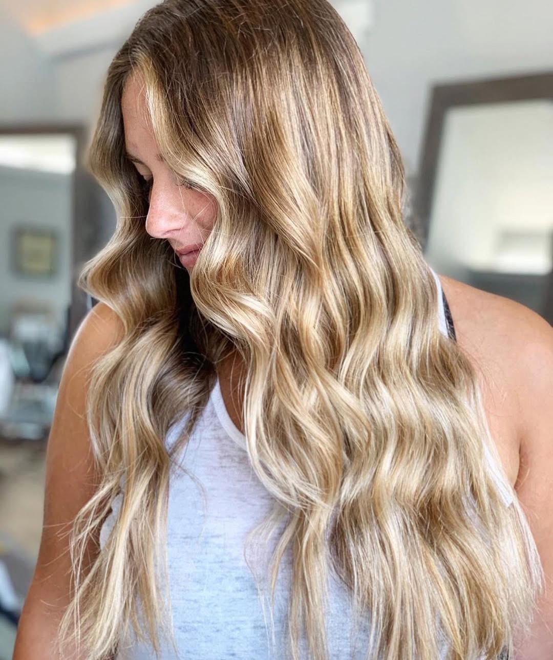 Close-up of a woman with long, sun-kissed blonde hair styled in soft, natural waves, perfect for summer beach hair ideas. She wears a flowy white dress against a blurred ocean background.