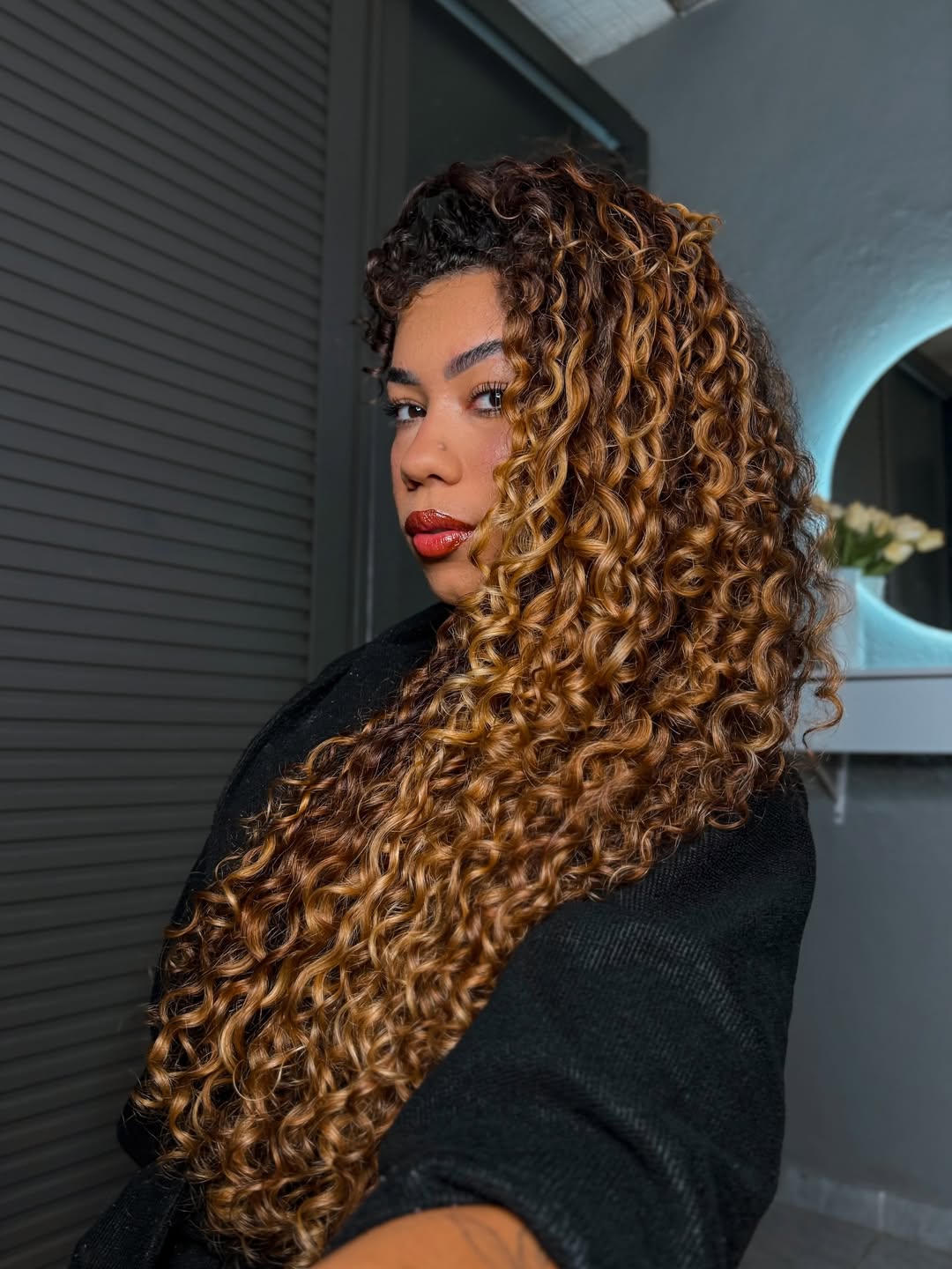 A woman with vibrant blonde summer curly hairstyles 2026, featuring beachy waves cascading over her shoulders. Her hair is perfectly styled, suggesting a sun-kissed, casual look for summer days.