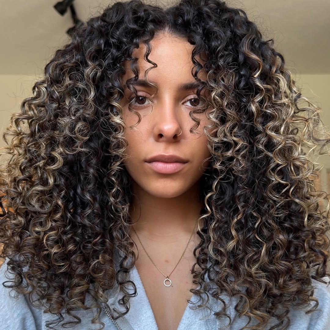 Beautiful woman smiling, showcasing her perfectly defined, bouncy summer curly hairstyles 2026. Her rich brown curls are styled with a side part, flowing gracefully over her shoulder, perfect for a chic daytime look.